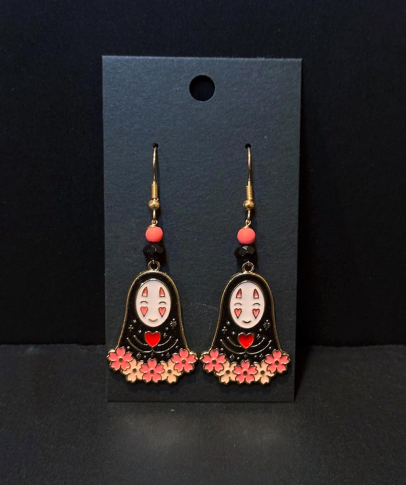 May include: A pair of gold-toned dangle earrings featuring a black figure with a white face and red heart. The earrings have pink flower accents and are suspended from gold hooks with pink and black beads. The earrings are displayed on a black card.