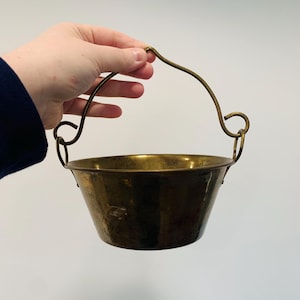 May include: A small, brass hanging planter with a simple, curved handle. The planter has a hammered finish and is perfect for displaying small plants or flowers.