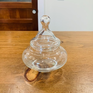May include: A clear glass jar with a lid. The jar has a rounded base and a lid with a small, rounded handle. The jar is sitting on a wooden surface. The jar is empty.