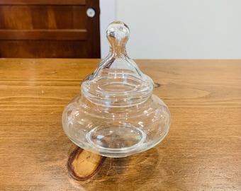 May include: A clear glass jar with a lid. The jar has a rounded base and a lid with a small, rounded handle. The jar is sitting on a wooden surface. The jar is empty.