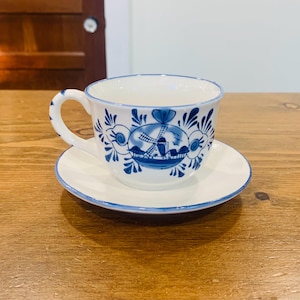 May include: White teacup and saucer set with a blue windmill and floral design. The cup has a curved handle and the saucer is round. The set is made of ceramic.