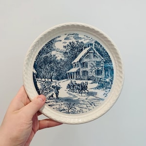 May include: A round, cream-colored plate with a blue and white transferware design. The scene depicts a winter landscape with a house, trees, and figures. The plate has a textured, rope-like border.