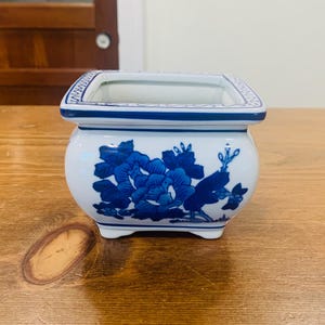 Op de afbeelding: Een vierkante keramische plantenbak met een witte basis en blauwe bloemmotieven. De rand is voorzien van een decoratieve blauwe geometrische rand. De plantenbak heeft een afgerond lichaam, een brede opening en vier kleine voetjes aan de onderkant.