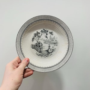 May include: A white ceramic plate with a black and white landscape design. The plate features a detailed illustration of a scenic view with a building, trees, and a person. The rim has a decorative black pattern.
