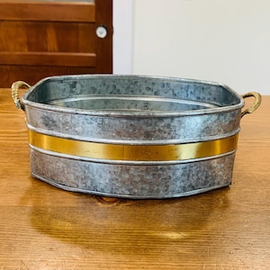 May include: A galvanized metal oval-shaped container with a gold band around the middle. The container has two rope-wrapped handles. The container is sitting on a wooden surface.
