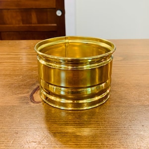 Op de afbeelding: Een cilindrische houder van gepolijst messing met een glad, reflecterend oppervlak en twee horizontale ribbels rondom het lichaam. De houder heeft een brede, open bovenkant met een licht uitlopende rand en een platte bodem in een goudkleur.
