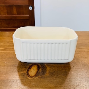 May include: A rectangular, off-white ceramic baking dish with vertical ribbed detailing on the sides. The dish has a smooth, rounded interior and sits on a wooden surface. The dish is empty and ready for use.