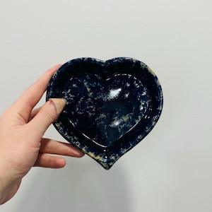 May include: A heart-shaped ceramic dish with a speckled navy blue glaze. The dish has a slightly raised rim and a smooth interior. The edges have a light tan color. The dish is held by a hand.