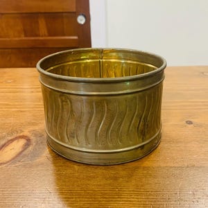 May include: A cylindrical brass container with a polished interior and a textured exterior featuring a repeating vertical wavy pattern. The metal vessel has a smooth rim and base, showcasing a warm metallic gold tone with subtle signs of age.
