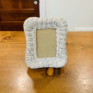 May include: A rectangular picture frame constructed from woven white wicker material. The frame features a thick, textured border with a braided pattern surrounding a plain, light tan rectangular insert in the center of the display area.