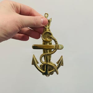 May include: A brass anchor door knocker with a rope design and a loop at the top for hanging. The anchor features a classic design with a crossbar and flukes. The knocker is held up against a white background.