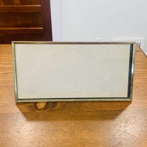 May include: A rectangular gold-colored picture frame with a decorative border. The frame has a light-colored backing and is displayed on a wooden surface. The frame is empty and ready for a photograph or artwork.