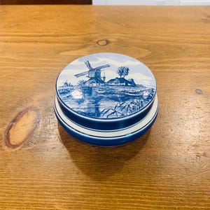 Może przedstawiać: Okrągłe, niebiesko-białe ceramiczne pudełko na drobiazgi. Pokrywka przedstawia szczegółową ilustrację wiatraka, drzew i wody w klasycznym niebiesko-białym schemacie kolorów. Pudełko ma wzór w paski po bokach.