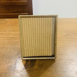 May include: A rectangular picture frame with a thin, metallic gold-toned border featuring a repeating geometric pattern. The interior contains a light beige, vertically ribbed paper insert that covers the majority of the display area.