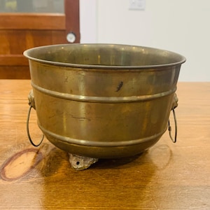 Könnte beinhalten: Eine runde Messingschale mit polierter Metalloberfläche, ausgestattet mit zwei seitlichen Griffen an Löwenkopfmotiven und drei kleinen Füßen an der Basis. Die Schale hat einen glatten, geschwungenen Körper mit horizontalen Rillen.