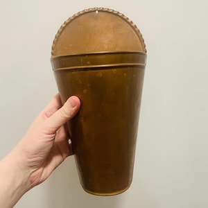 Op de afbeelding: Een koperen wandplantenbak met een halfronde bovenkant en een taps toelopende, conische vorm. Het metalen oppervlak heeft een warme, bruine kleur met subtiele kleurvariaties en een geschulpte rand aan de bovenkant voor wandmontage.