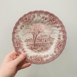 May include: A decorative, vintage-style plate with a detailed landscape scene in shades of red and white. The plate features a rural scene with a house, bridge, and tree. The rim is adorned with floral and vine patterns.