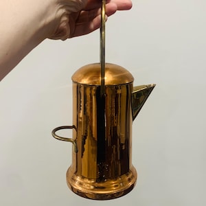 May include: A copper-coloured vintage tea kettle with a brass handle and spout. The kettle has a cylindrical body with a rounded base and a lid. The handle is arched, and the spout is triangular. The kettle is held by a hand.