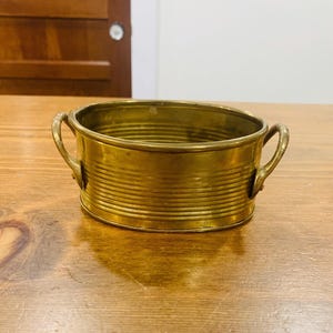 May include: A small, antique brass planter with a horizontal ribbed design. The oval-shaped container has two curved handles on either side. The planter is sitting on a wooden surface.