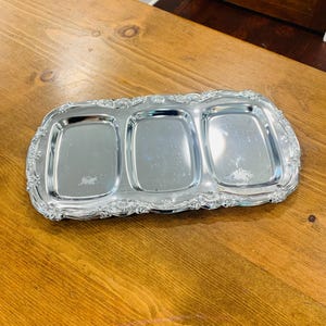 May include: A rectangular silver-toned metal serving tray featuring three distinct recessed compartments. The tray has a polished, reflective surface and is bordered by an ornate, raised decorative rim with intricate scrollwork detailing.
