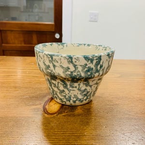 Op de afbeelding: Een keramische bloempot met een crèmekleurige binnenkant en een gespikkelde teal en crèmekleurige buitenkant. De pot heeft een klassieke, licht uitlopende vorm en staat op een houten oppervlak. De pot is leeg en klaar om te planten.