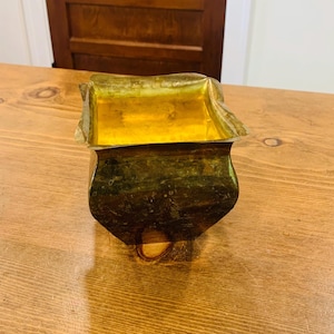 May include: A square-shaped brass container with a weathered, dark patina on the exterior sides and a bright, polished gold-toned interior. The top rim features a distinct wavy, scalloped edge design that curves outward at each of the four corners.