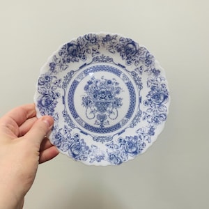 May include: A white ceramic plate with a scalloped edge featuring a blue floral pattern. The design includes a central bouquet of flowers surrounded by a decorative lattice border and a rim adorned with detailed roses and foliage in blue ink.