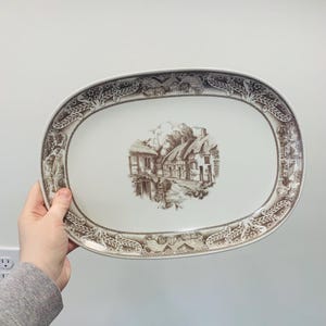 May include: Oval serving platter with a brown and white transferware design. The center features a detailed illustration of a village scene. The rim is decorated with floral and architectural motifs. A classic piece for serving food.