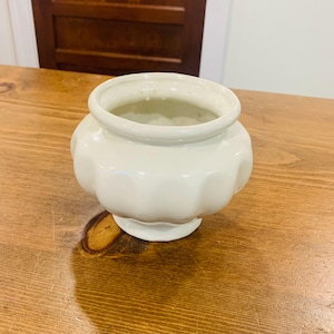 May include: A small, off-white ceramic bowl with a rounded, bulbous body and a wide, circular opening. The exterior features a series of vertical, scalloped indentations around the midsection, resting on a short, flared pedestal base.