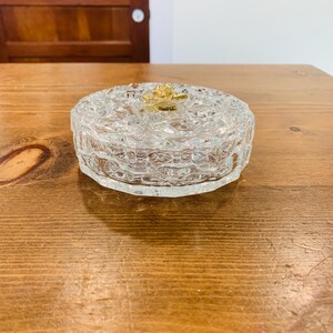 May include: A clear glass trinket box with a gold rose design on the lid. The box has a textured, patterned surface.