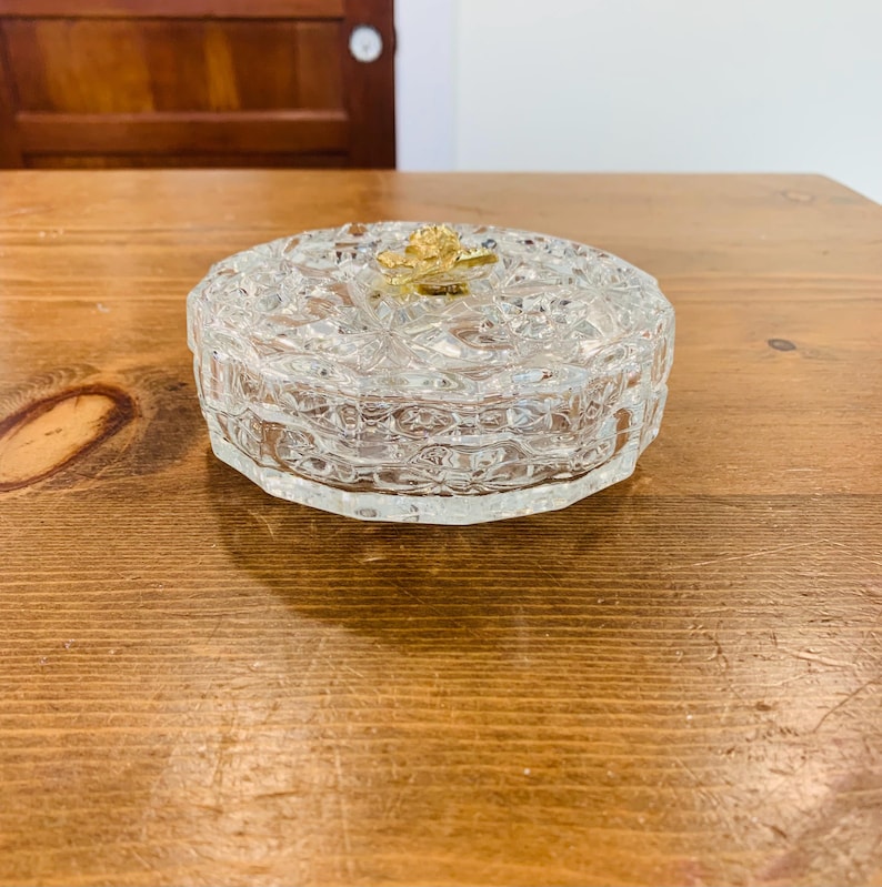 May include: A clear glass trinket box with a gold rose design on the lid. The box has a textured, patterned surface.