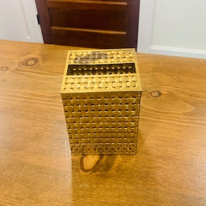 May include: A square tissue box cover with a metallic gold finish and a woven basket-weave texture. The top features a rectangular opening for dispensing tissues, and the base is accented with a decorative band of repeating triangular patterns.