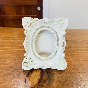 Peut inclure: Un petit cadre photo en céramique blanc cassé avec une ouverture ovale. Le cadre présente un design orné et décoratif avec un aspect légèrement vieilli et patiné. Le cadre est posé sur une surface en bois.