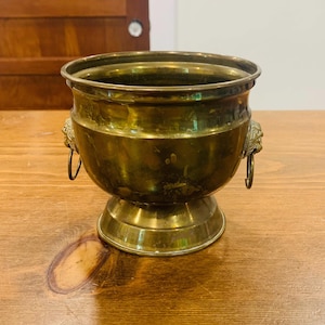 Pode incluir: Uma taça de latão polido com uma borda larga e uma base de pedestal. O exterior apresenta duas alças decorativas em forma de cabeça de leão nos lados opostos, cada uma segurando um pequeno anel circular. A superfície metálica é dourada.