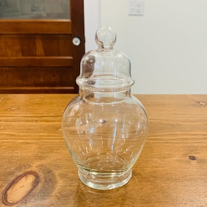 May include: A clear glass apothecary jar with a rounded body and a matching removable lid featuring a spherical knob handle. The container has a wide base that tapers upward to a narrow neck before widening into a bulbous shape, topped by a lid.
