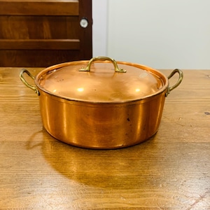 Könnte beinhalten: Ein Kochtopf aus Kupfer mit Deckel und Messinggriffen. Der Topf ist rund mit einer glänzenden, reflektierenden Oberfläche. Der Deckel hat einen Messinggriff. Der Topf steht auf einem Holztisch.
