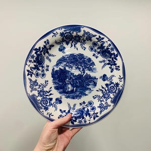 May include: A round ceramic plate featuring a white base with a deep blue transferware pattern. The center depicts a landscape scene with figures near a tree and bridge, while the rim is decorated with a dense floral motif of blossoms and leaves.