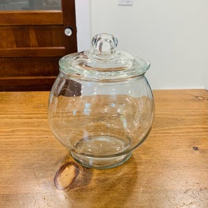 Op de afbeelding: Een doorzichtige glazen voorraadpot met een ronde, bolvormige behuizing en een bijpassend afneembaar glazen deksel. Het deksel is voorzien van een kleine, bolvormige knop als handvat aan de bovenzijde. De pot heeft een brede opening en een platte voet.