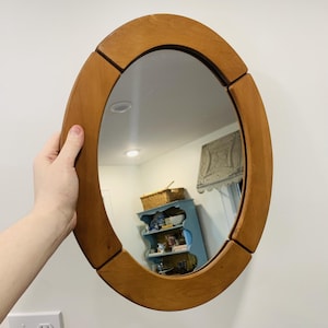 May include: An oval-shaped mirror featuring a smooth, light brown wooden frame constructed from four distinct, curved segments. The reflective glass surface is clear and shows a reflection of an interior room with a blue shelf and a basket.