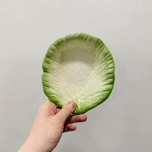 May include: A ceramic bowl shaped like a cabbage leaf with a white center and vibrant green edges. The surface features a detailed, embossed texture that mimics the natural veins and ruffled appearance of a real vegetable leaf in a circular form.