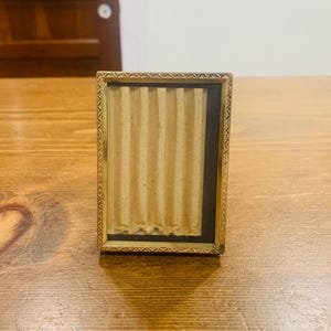 May include: A rectangular picture frame with a metallic gold finish and a textured, repeating triangular pattern along the border. The interior features a vertical, pleated beige paper insert set against a dark black background.