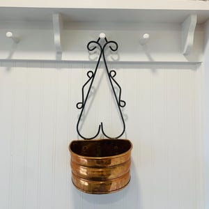 May include: A wall-mounted copper planter with a black wrought iron frame. The planter has a three-tiered design and is suspended from a decorative, scrolled bracket. The copper has a warm, aged patina, adding to its vintage appeal.