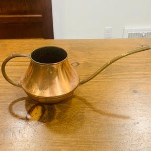 May include: A copper watering can with a wide, rounded base, a curved handle on the side, and a long, slender, tapered spout extending outward. The metal surface shows a warm, metallic finish with subtle signs of natural oxidation and wear.