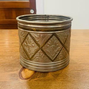 Könnte beinhalten: Ein zylindrischer Metallbehälter mit silberfarbenem Finish und einem erhabenen Rautenmuster auf der Außenseite. Die Oberfläche weist geprägte florale Motive innerhalb jeder Rautenform auf, akzentuiert durch dunkle, verwitterte Details.