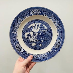 May include: A blue and white Willow pattern plate. The plate features a central scene with a pagoda, birds, and a bridge, surrounded by a detailed border. The plate is held by a hand against a white background.