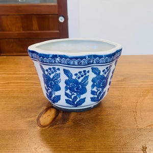 Könnte beinhalten: Ein weiß-blauer Keramik-Blumentopf mit floralem Design. Der Topf hat einen gewellten Rand und eine Reihe blauer Blumenmuster an den Seiten. Der Topf steht auf einer Holzoberfläche.