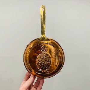 May include: A round copper pan featuring a raised pineapple design embossed in the center. The pan has a long, flat brass handle attached with two rivets at the base. The metallic surface is polished and reflective, showing a warm orange-brown hue.