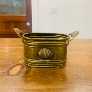 May include: An oval brass planter featuring a horizontal ribbed design and two side handles. The front of the vessel is decorated with a centered, embossed seashell motif. The metal has a warm, aged patina with visible golden tones throughout.