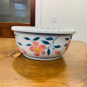 Op de afbeelding: Witte keramische kom met een geschulpte rand, versierd met roze bloemen, blauwgroene bladeren en blauwe accenten. Het bloemmotief is met de hand geschilderd op de buitenkant van de kom. Deze decoratieve kom is geschikt om te serveren of tentoon te stellen.