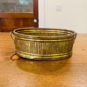 May include: An antique brass oval planter with vertical ribbed detailing. The planter has two small handles on either side and sits on a wooden surface. The brass has a warm, aged patina.